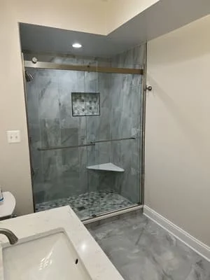 Frameless glass shower with gray marble tile walls, pebble stone floor, and built-in bench