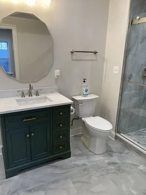 Dark green vanity with marble countertop, marble tile floor, frameless glass shower, and round mirror in renovated bathroom